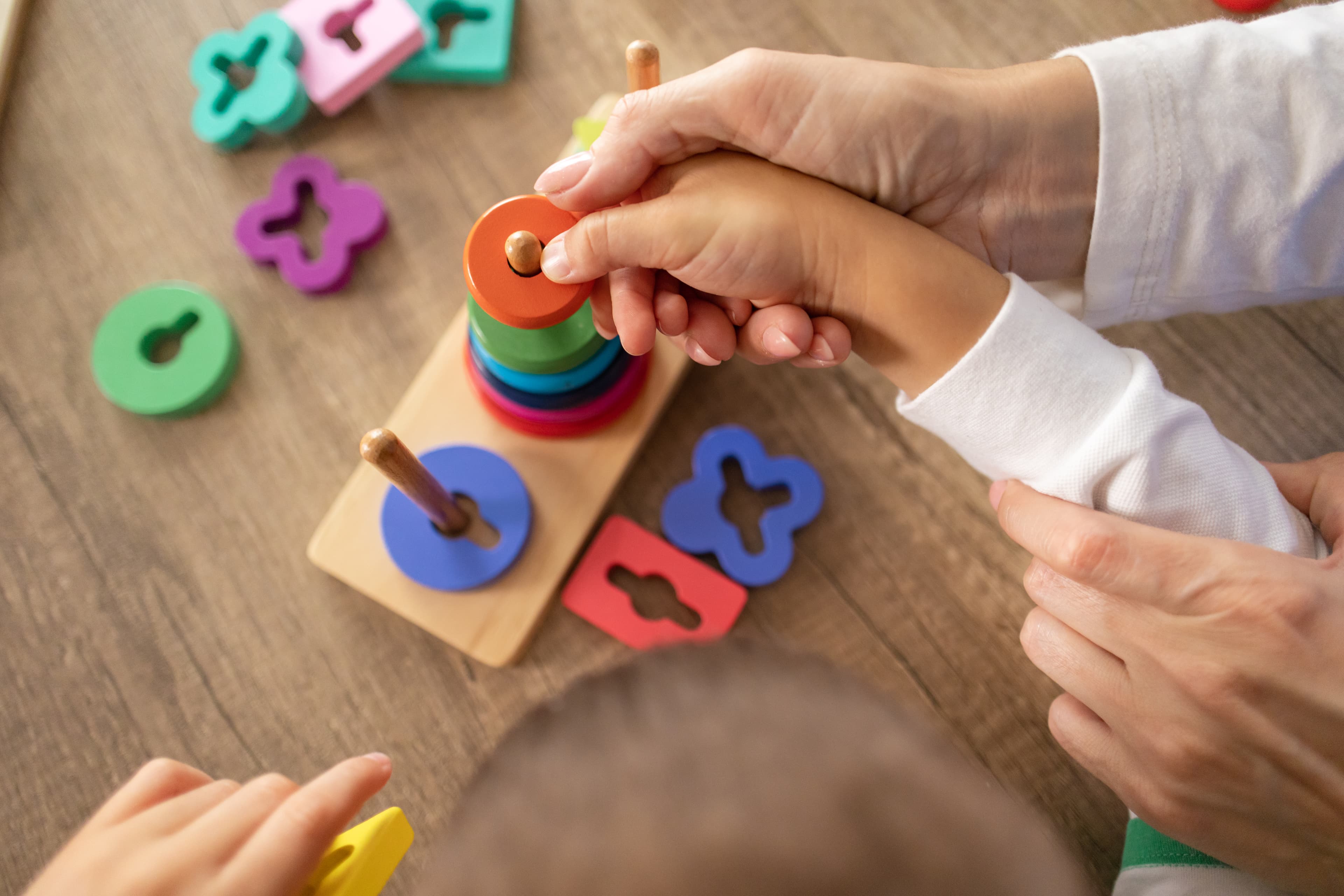 Adult hands guiding a child's hand to stack colorful wooden rings on a peg.