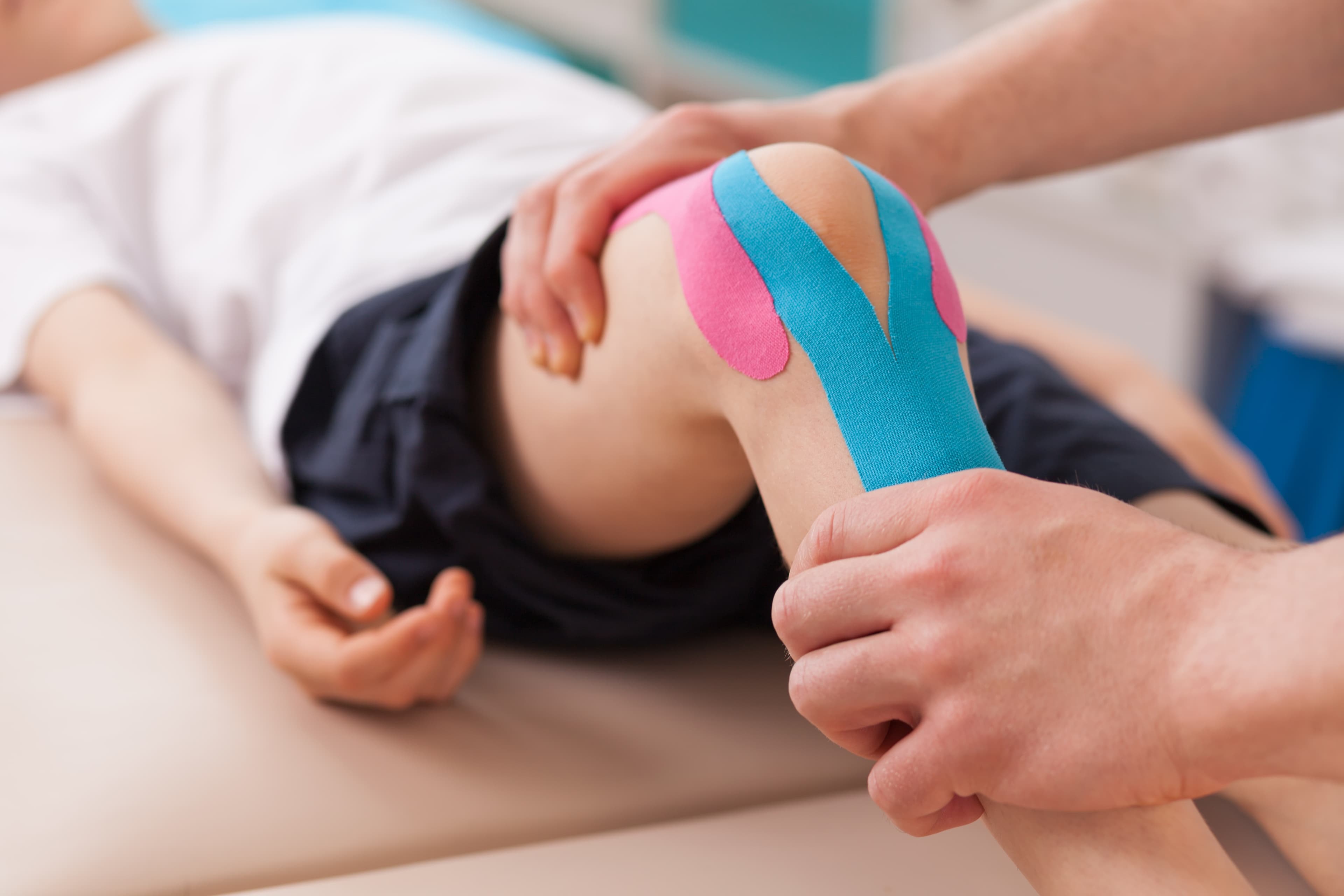 Physical therapist applying pink and blue kinesiology tape to a patient's knee for injury support.